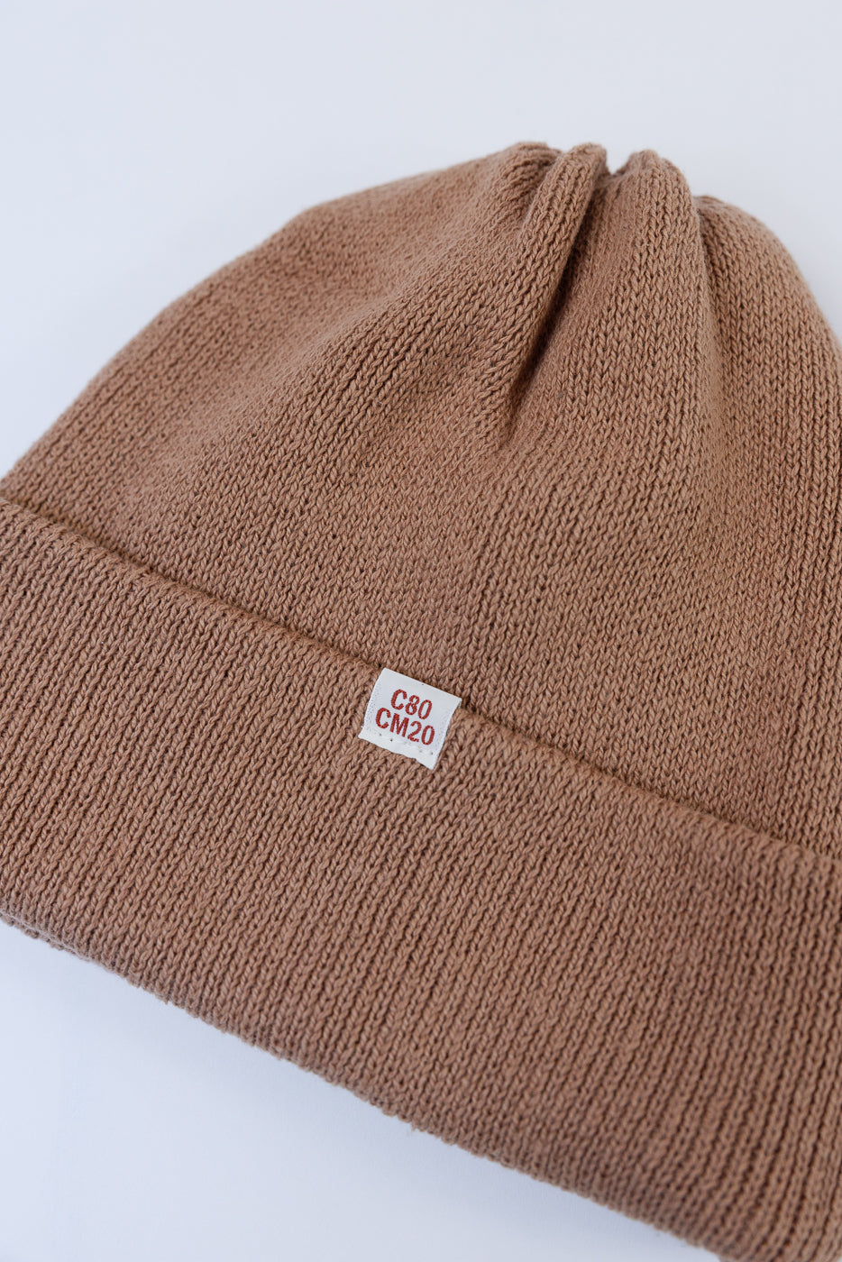 Close-up front view of a tan knit beanie showing fabric texture and a small white tag with red text.
