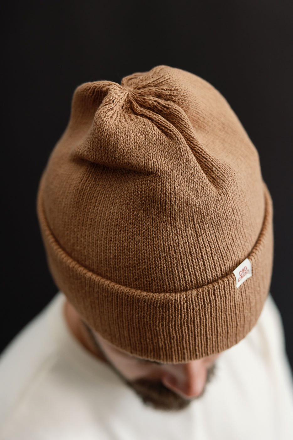 Close-up front view of a brown knit beanie on a male model showing fabric texture and tag.