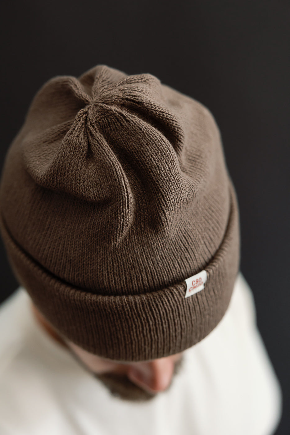 Close-up front view of a brown ribbed knit beanie with a small white brand tag.