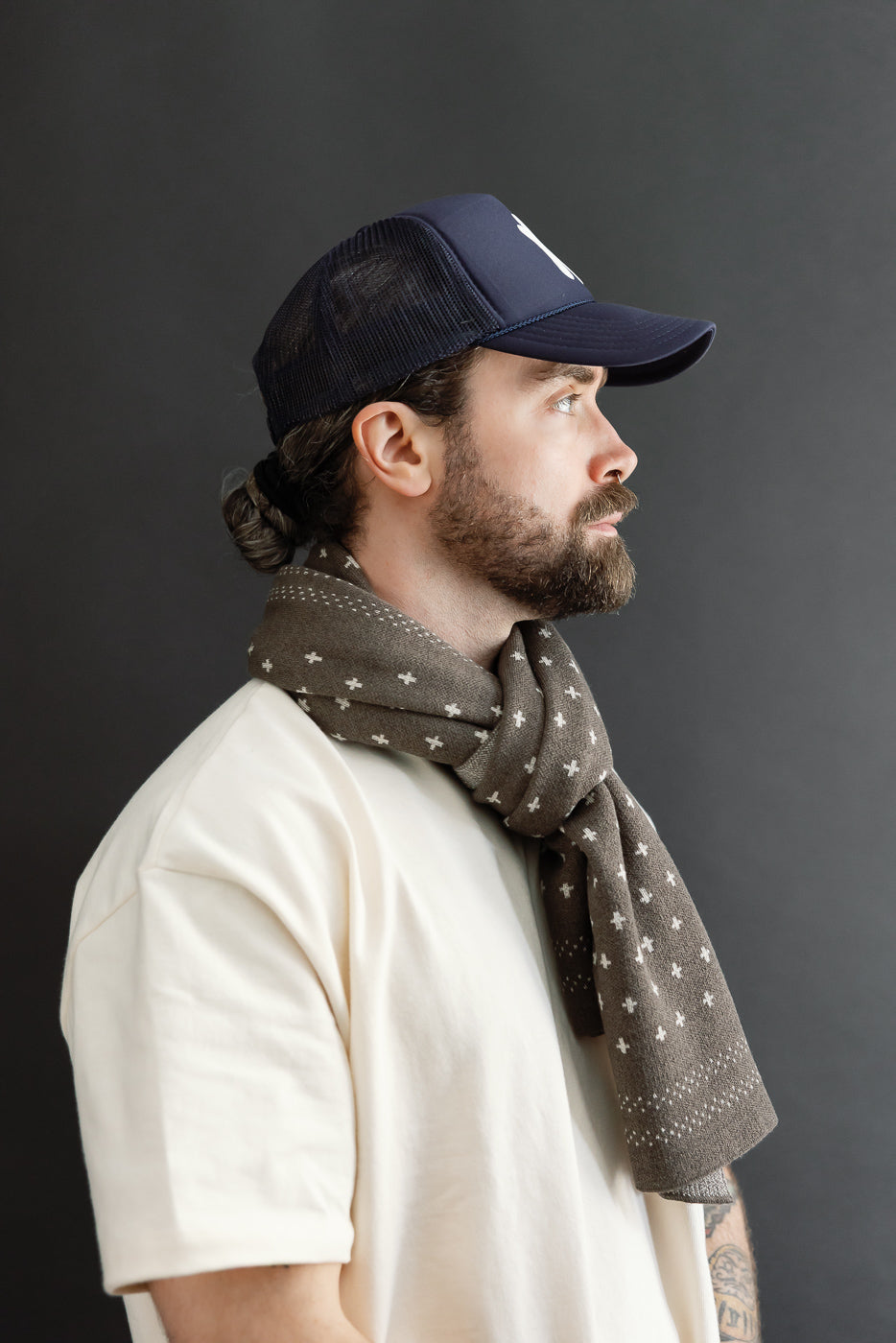 Side profile close-up of male model wearing a navy trucker hat and patterned brown scarf.