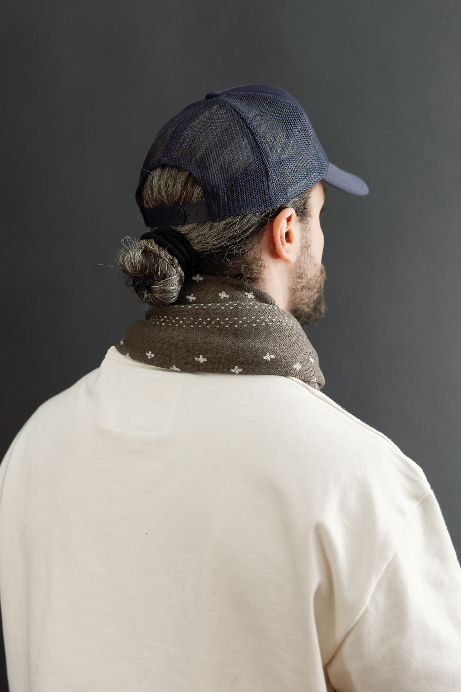 Back view close-up of a male model wearing a navy mesh trucker hat and a patterned brown neck scarf.
