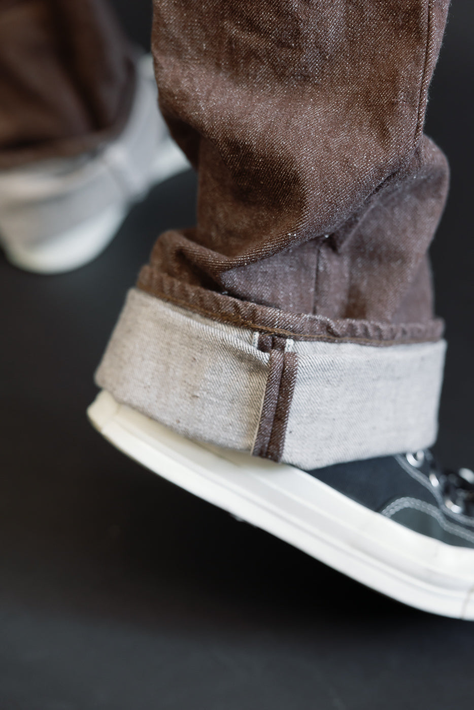 Close-up of brown selvedge jeans hem, showing cuffed detail and texture, paired with sneakers.
