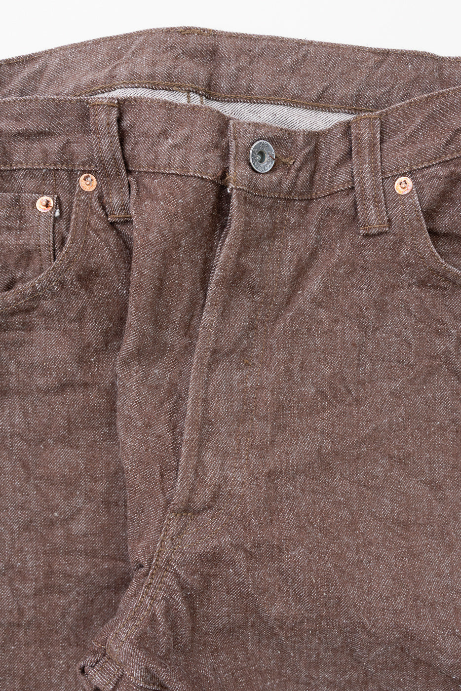 Close-up front waist of brown selvedge denim jeans, showing copper rivets, button fly, and textured fabric weave