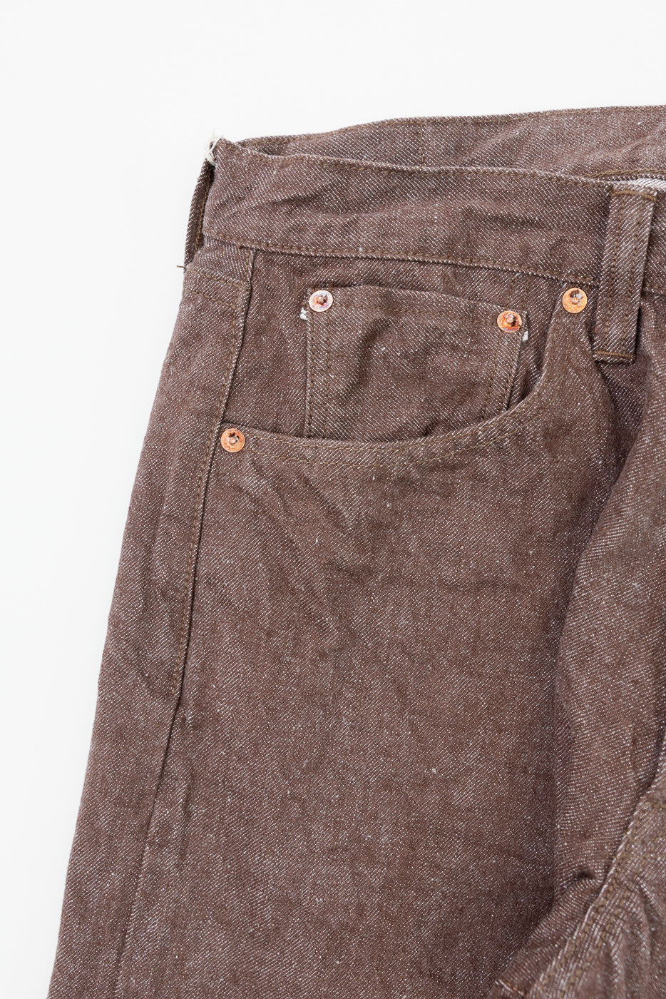 Close-up front view of brown selvedge denim jeans, copper rivets, textured fabric, front pocket detail