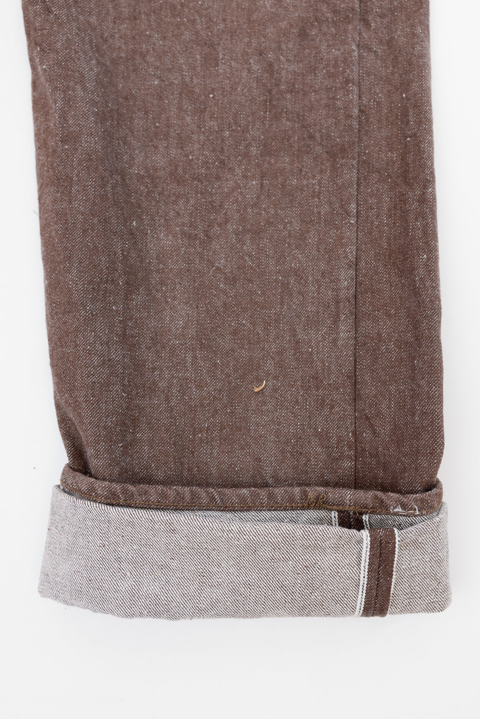 Close-up of brown selvedge denim pant leg, front view, showing cuffed hem with contrast stitching and fabric texture