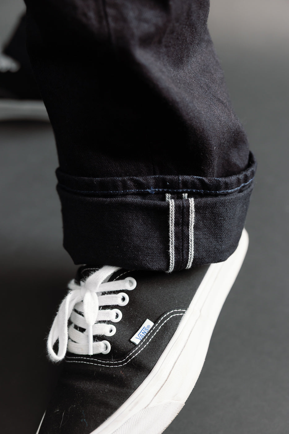 Close-up of Samurai S0510XX Aoki Tachikage black blue selvedge denim leg, hem, and side seam detail, with cuffed hem above black Vans sneaker; left pant leg shown.