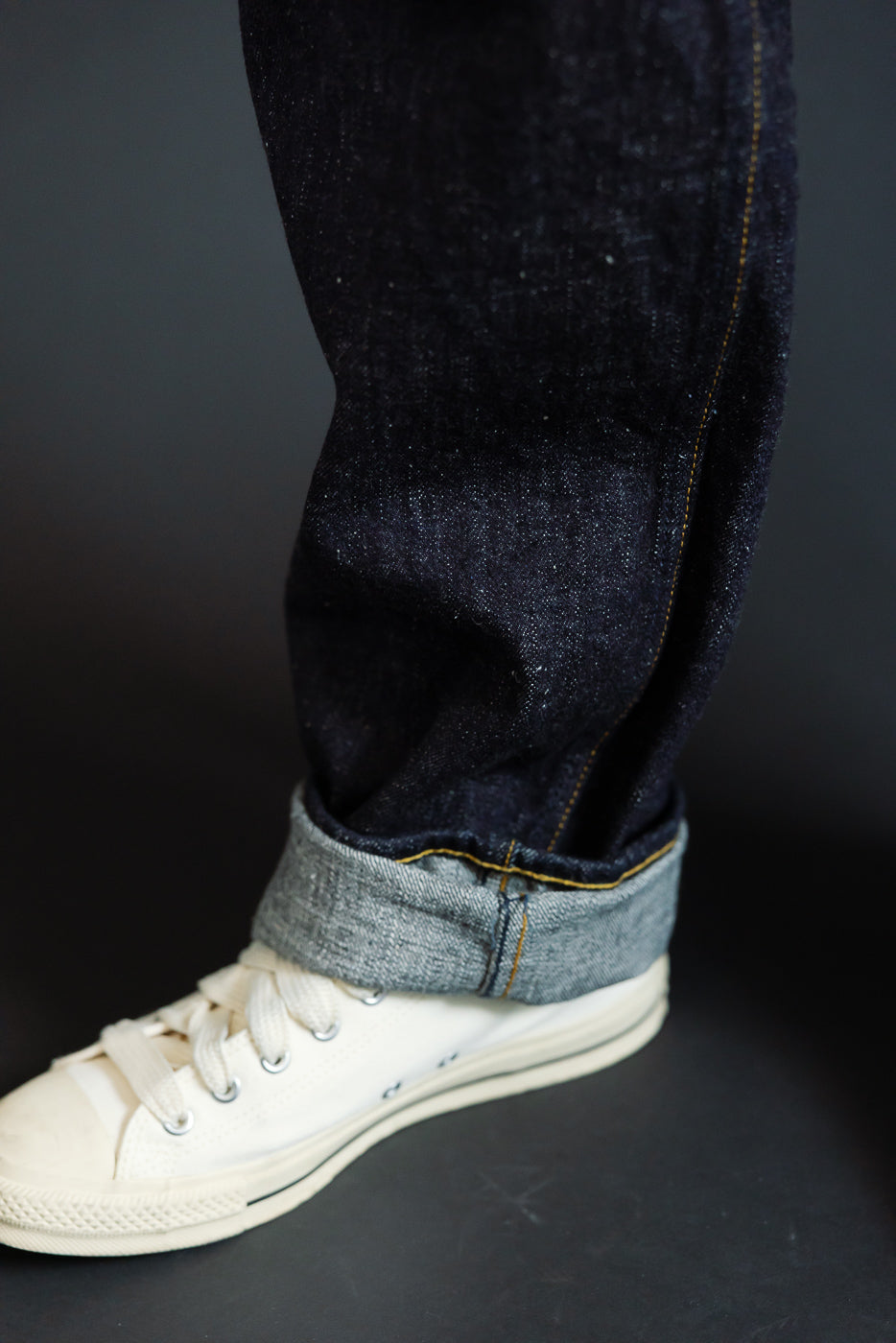 Close-up side view of male model's leg in dark selvedge denim jeans with cuffed hem, showing raw edge and gold stitching, paired with white sneakers