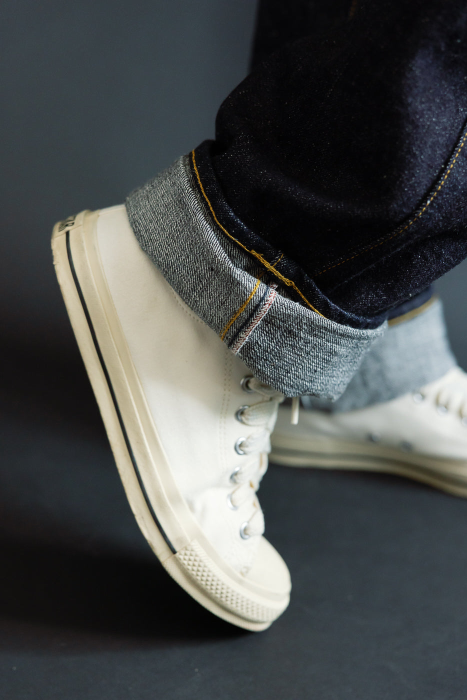 Close-up side view of male model's cuffed selvedge denim jeans with yellow stitching, paired with off-white high-top sneakers