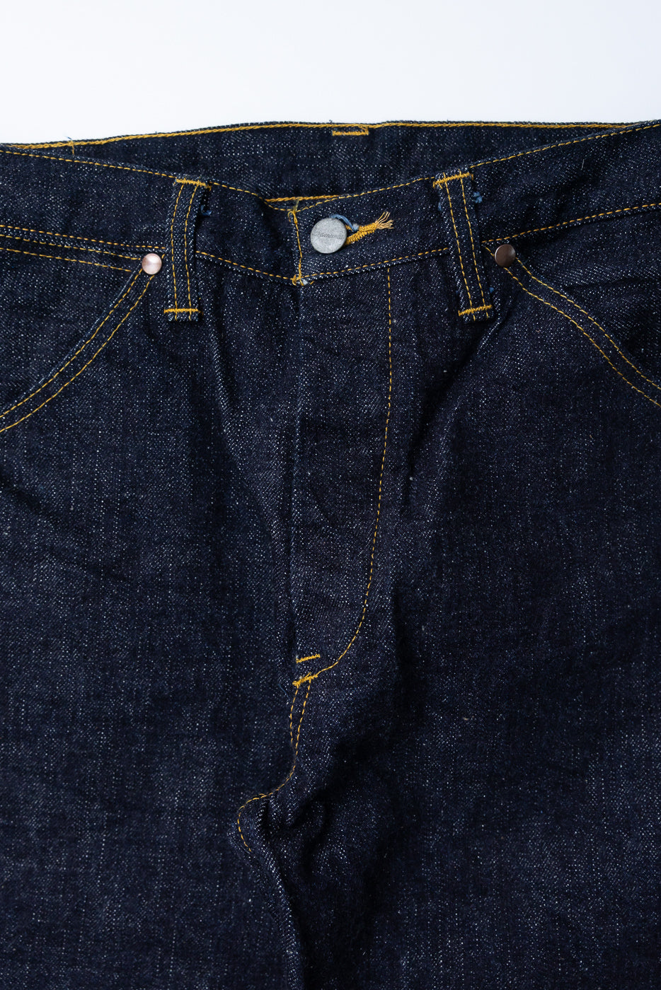 Close-up front view of raw selvedge denim jeans showing button fly, yellow stitching, and metal rivets on dark indigo fabric.