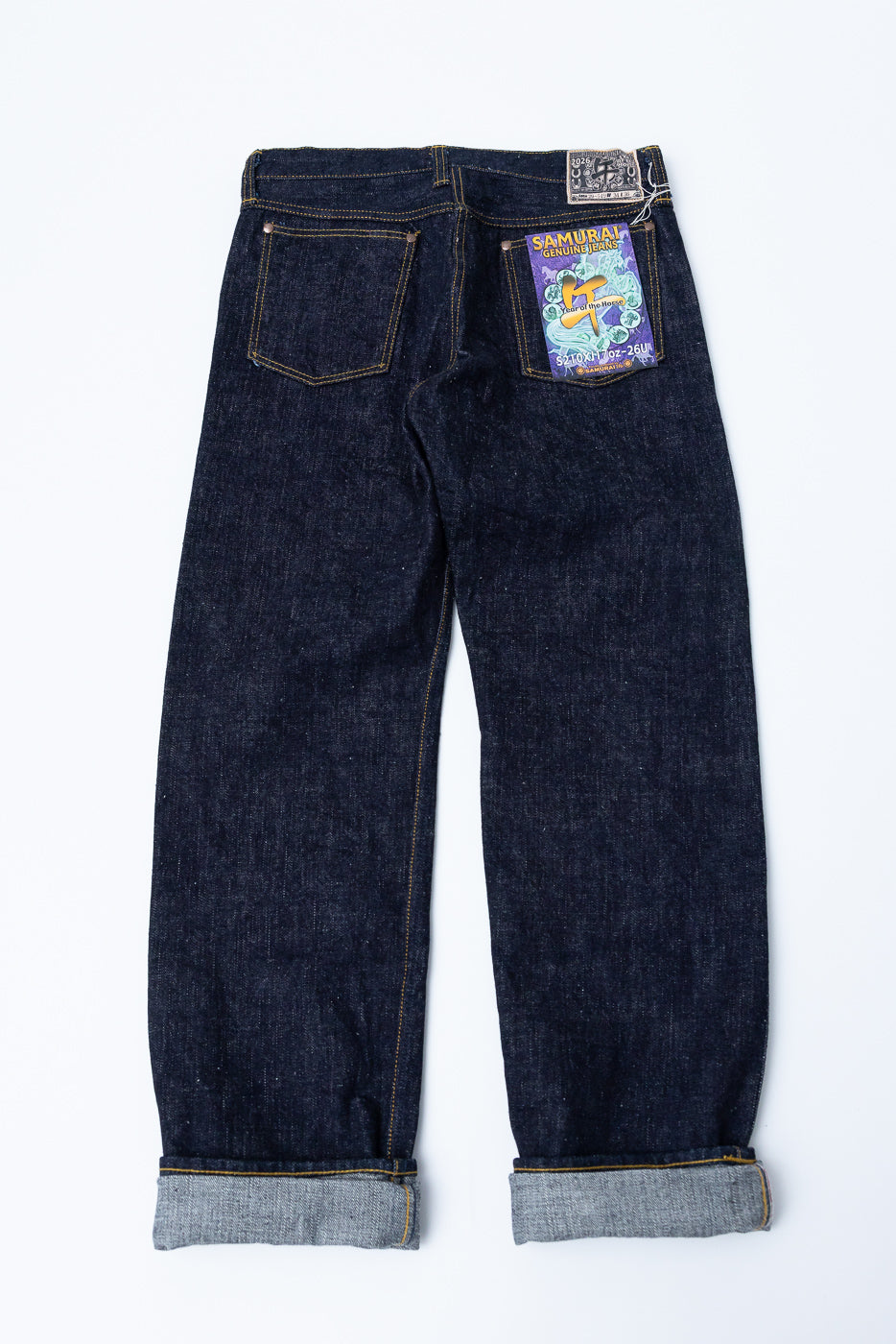 Back view flat lay of dark selvedge denim jeans, showing back pockets, waistband tag, and cuffed hems with contrast stitching.