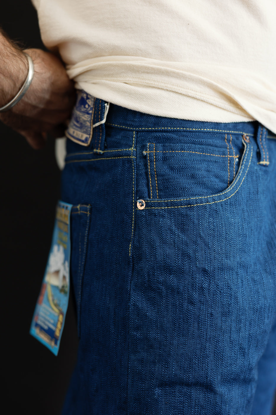 Close-up side view of men's selvedge denim jeans, showing back pocket, copper rivet, yellow stitching, and fabric texture on athletic build model.