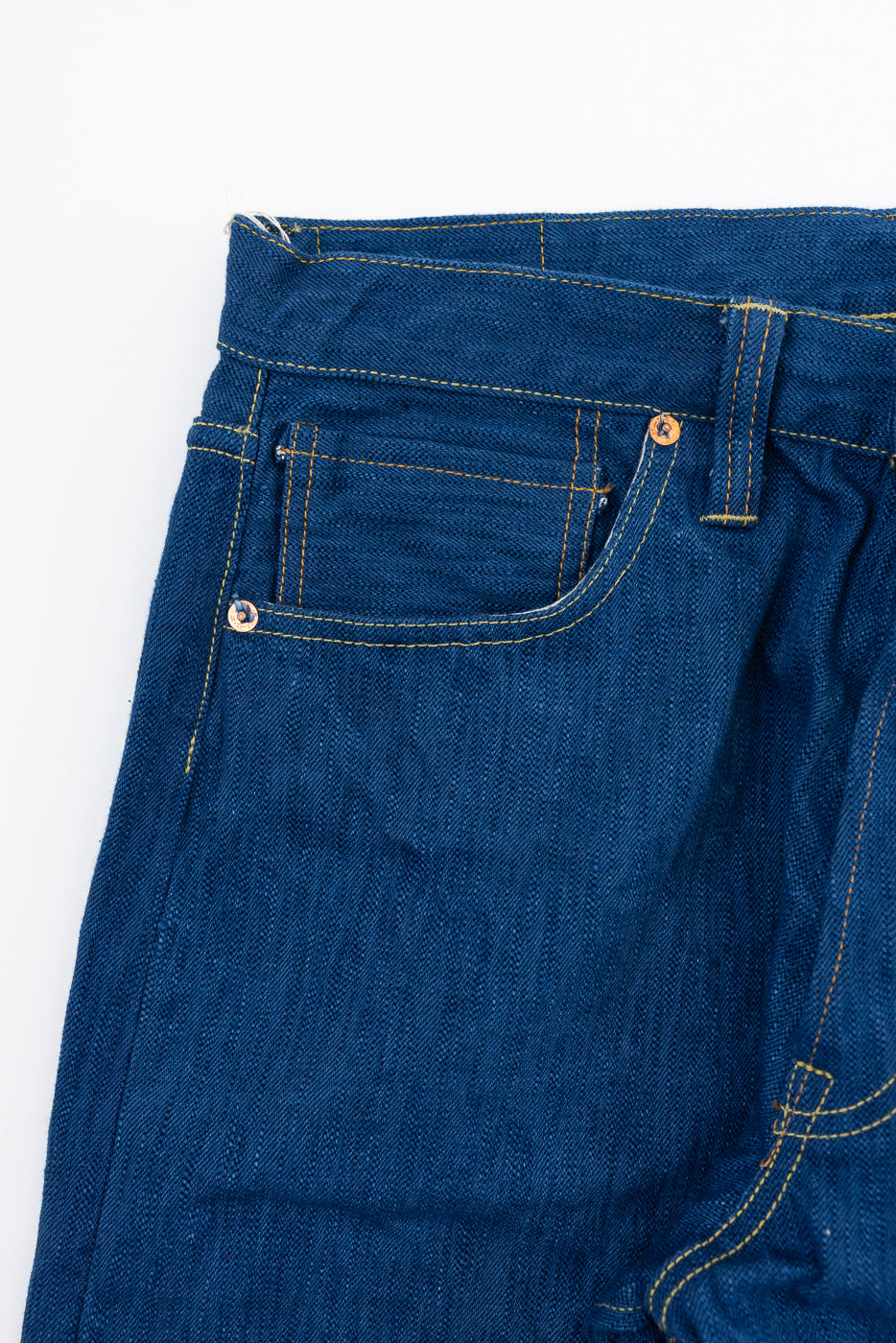 Close-up front view of raw selvedge denim jeans showing coin pocket, copper rivets, and yellow stitching detail