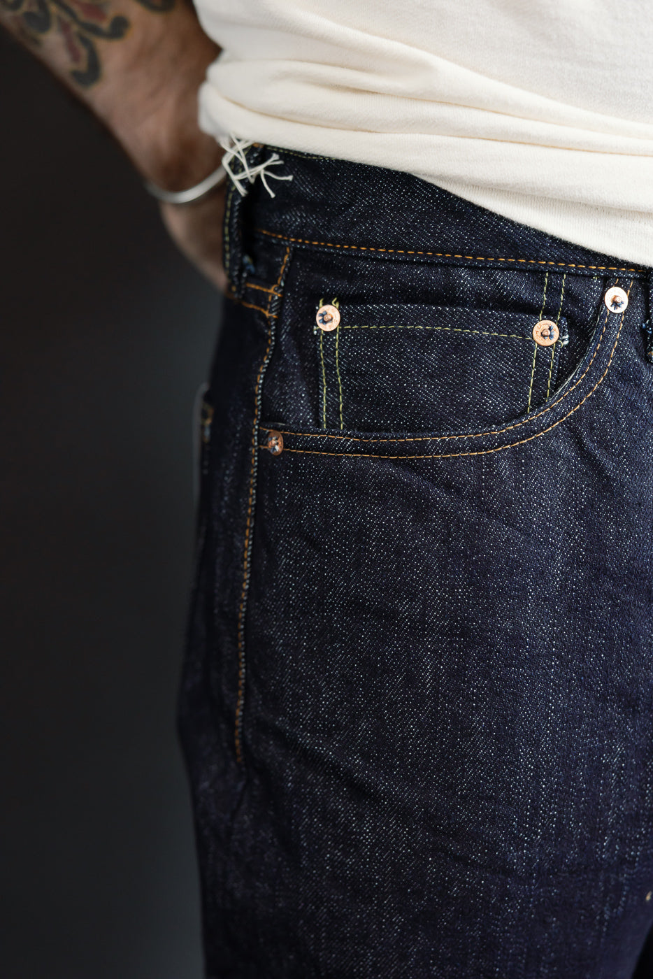 Close-up front view of dark selvedge denim jeans on male model, showing copper rivets, contrast stitching, and pocket details