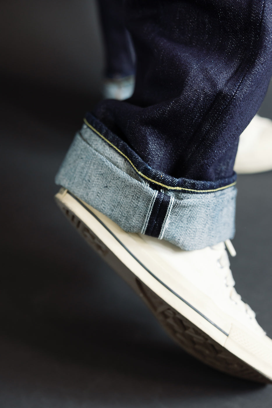 Close-up side view of male model’s cuffed selvedge denim hem with yellow stitching, paired with white sneakers