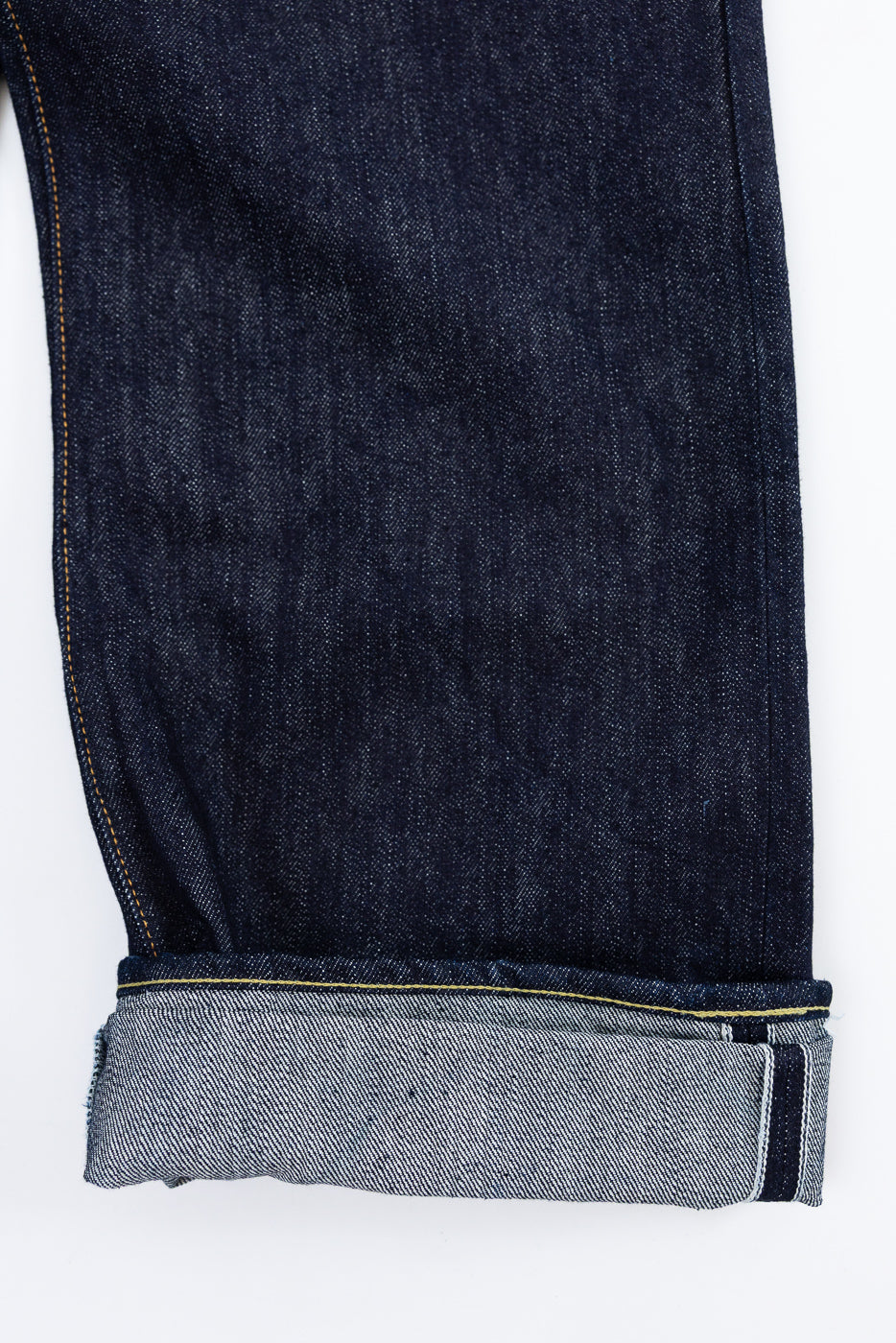 Close-up front view of dark selvedge denim jeans leg, showcasing raw hem, contrast stitching, and fabric texture