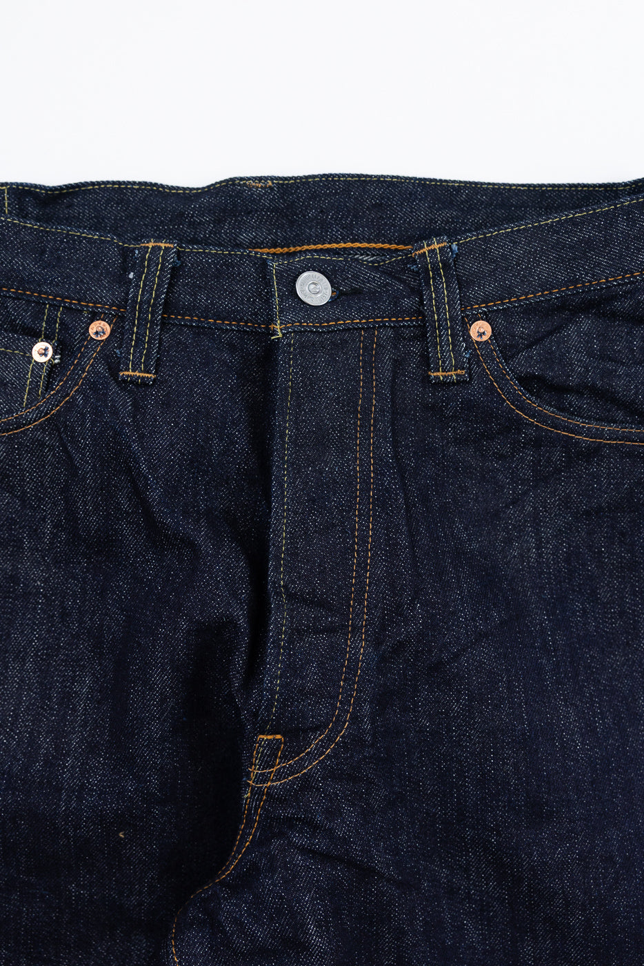 Close-up front view of raw selvedge denim jeans showing button fly, copper rivets, and contrast stitching.