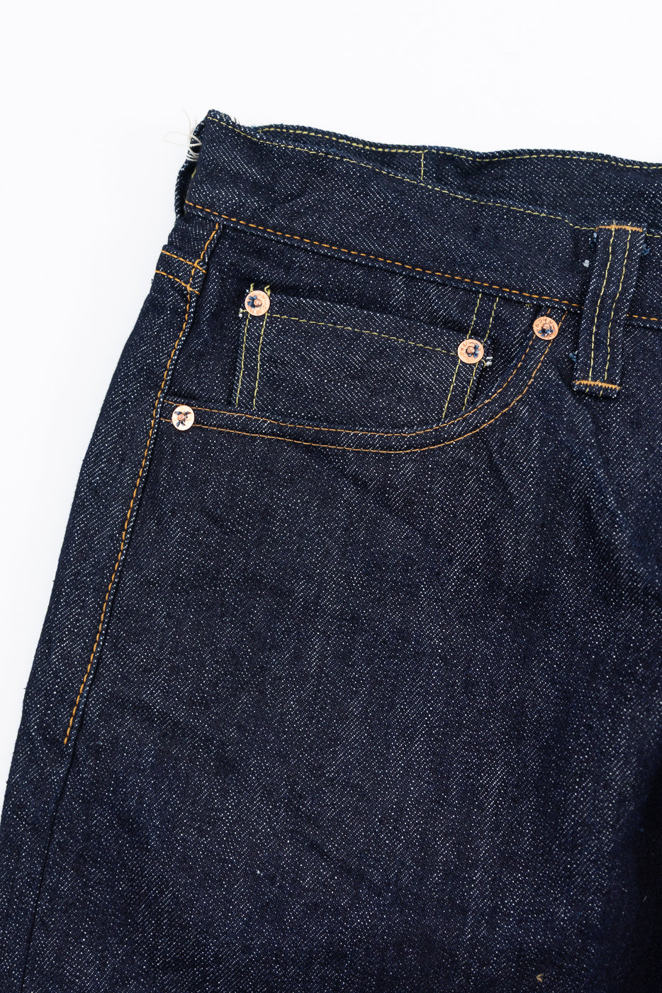 Close-up front view of men's selvedge denim jeans, showing copper rivets, orange stitching, and raw hem detail.