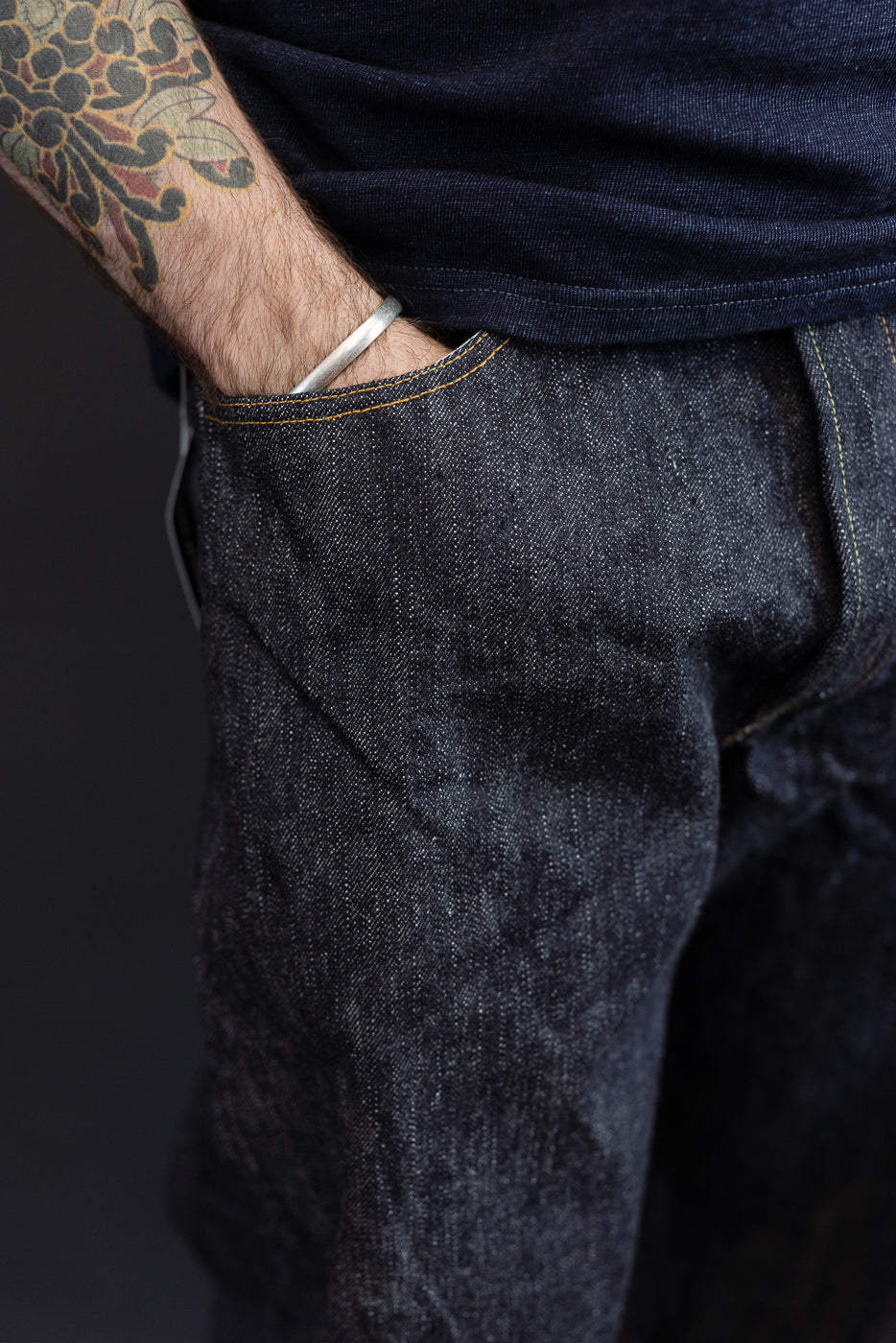 Close-up side view of dark selvedge denim jeans on male model, showing front pocket, orange stitching, and fabric texture