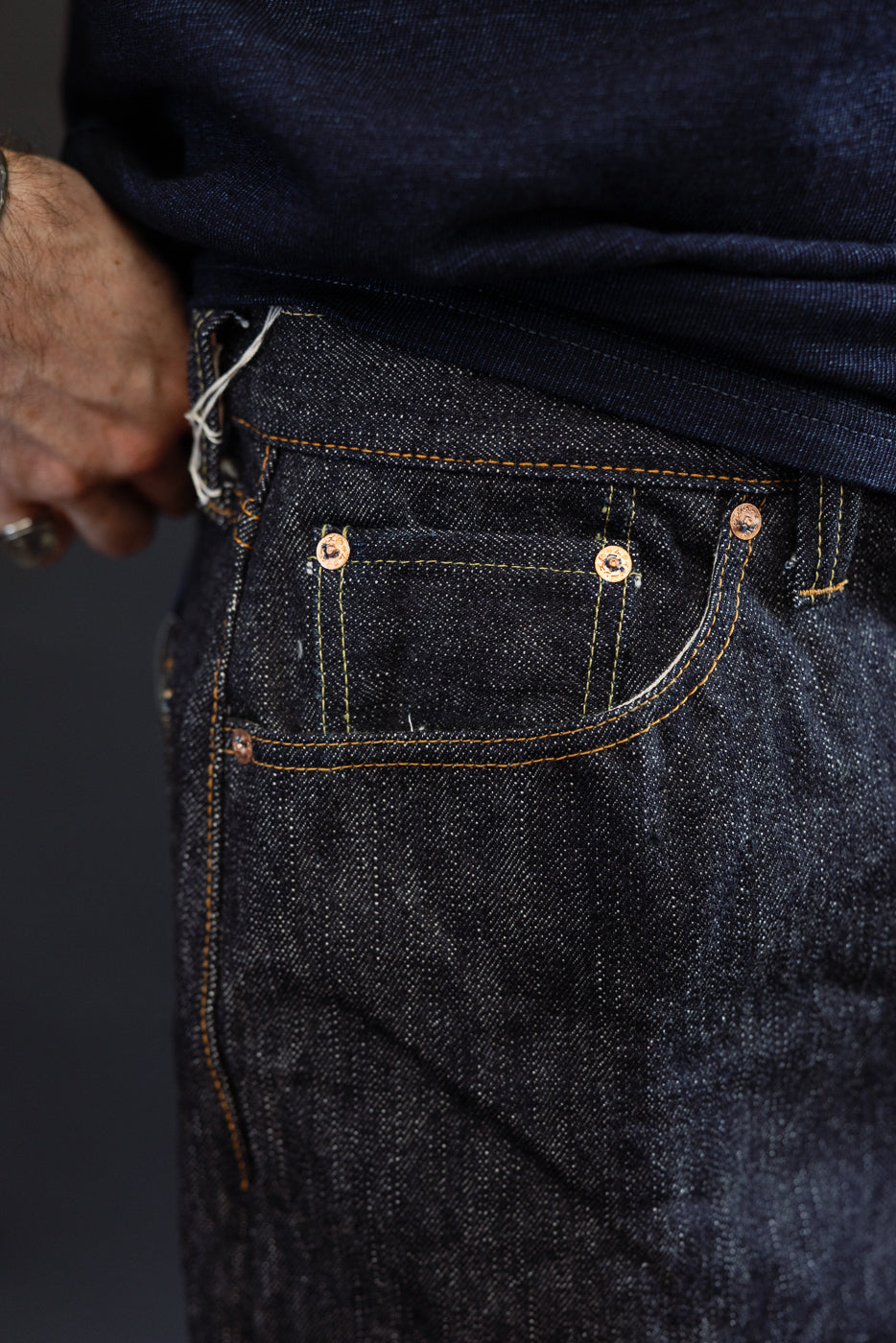 Close-up front view of men's dark selvedge denim jeans, showing coin pocket with copper rivets, orange stitching, and fabric texture on athletic build model.