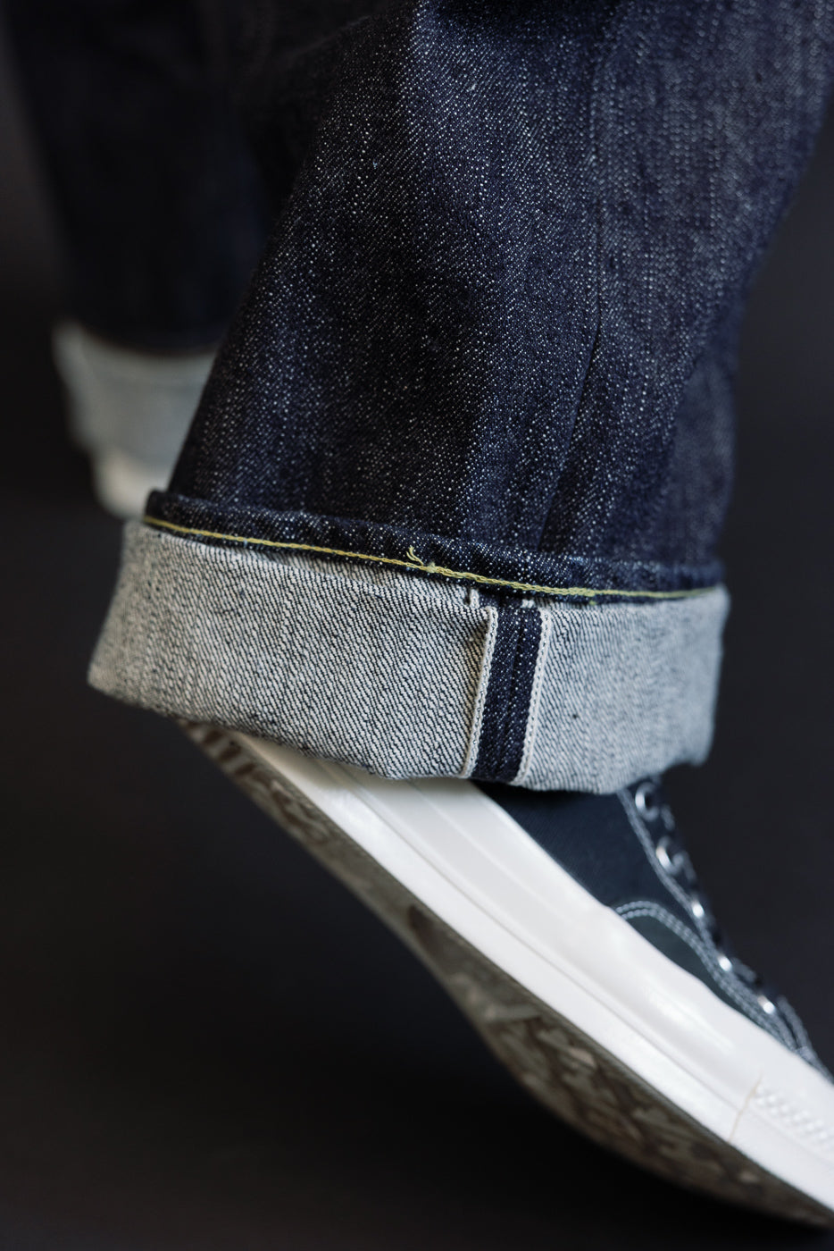 Close-up side view of dark selvedge denim jeans cuff, showing raw hem, yellow stitching, and fabric texture on male model.