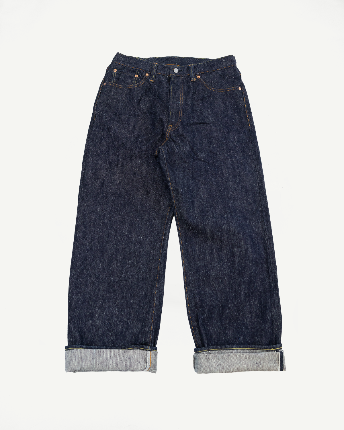 Front view flat lay of dark selvedge denim jeans with cuffed hems showing raw edge, copper rivets, and contrast stitching