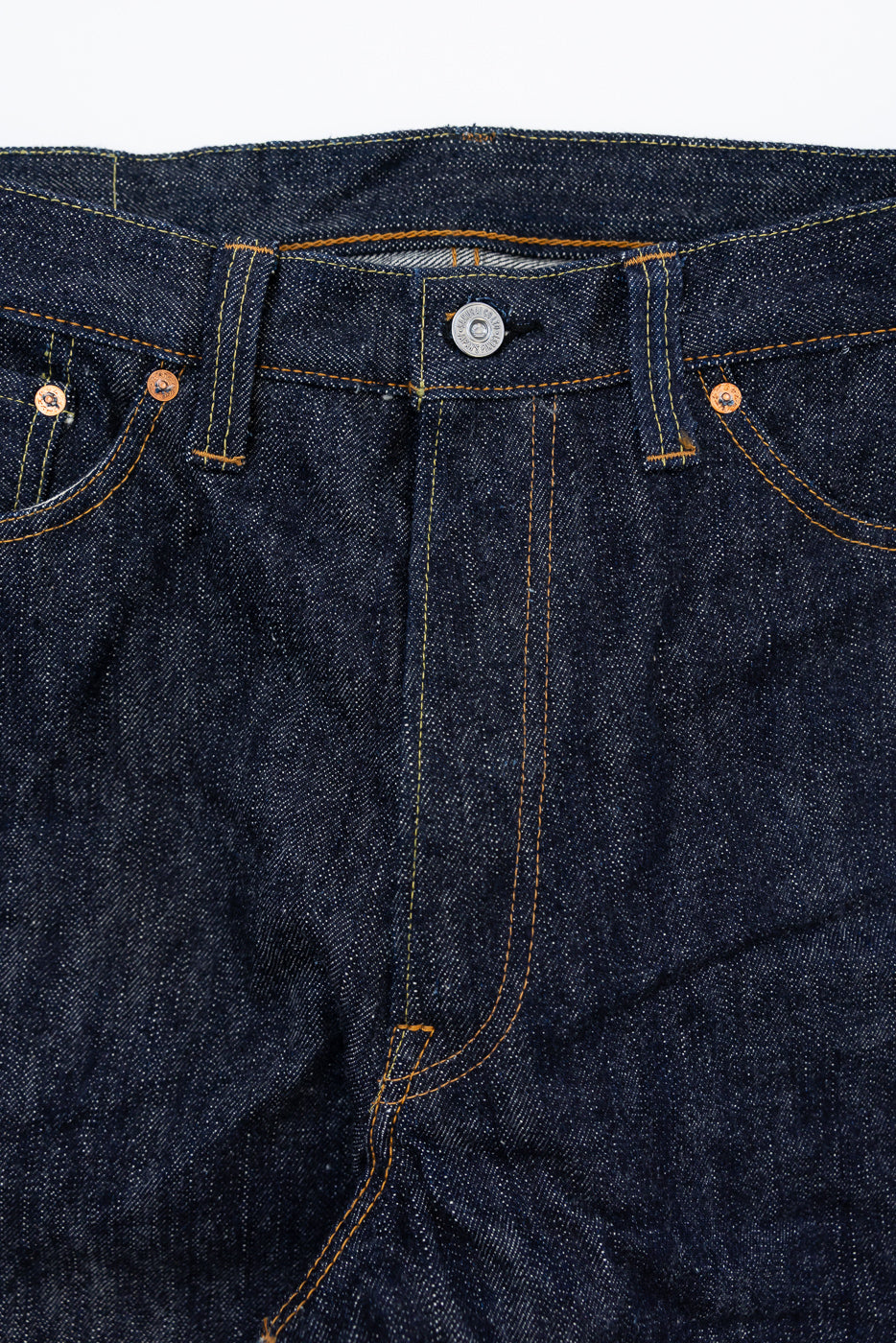 Close-up front of raw selvedge denim jeans showing button fly, copper rivets, and orange stitching detail