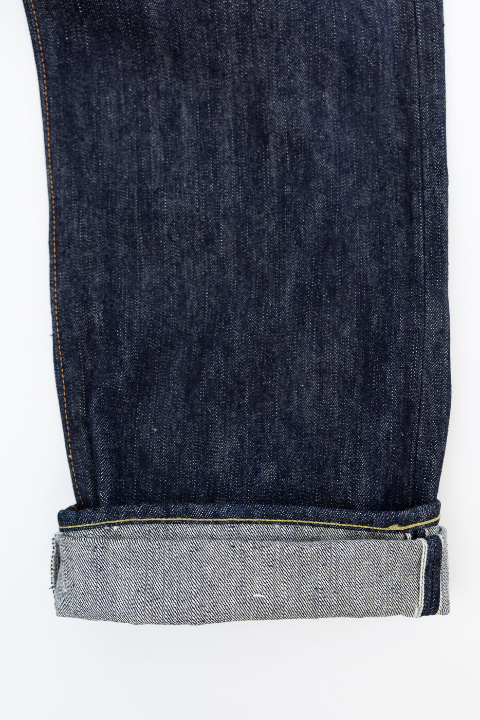 Close-up front view of raw selvedge denim jeans leg, showcasing dark indigo fabric, yellow stitching, and cuffed hem with selvedge edge.