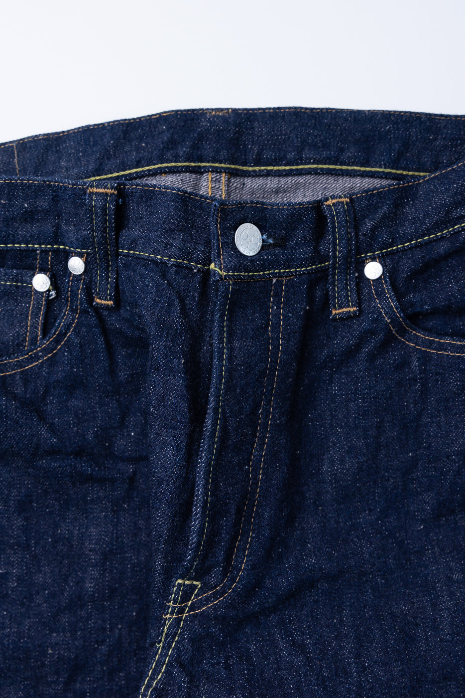 Close-up of Samurai's S500AX-SAKURA jeans, showcasing the dark blue denim waistband, sakura motif button, rivets, belt loops, and distinctive yellow-orange topstitching.