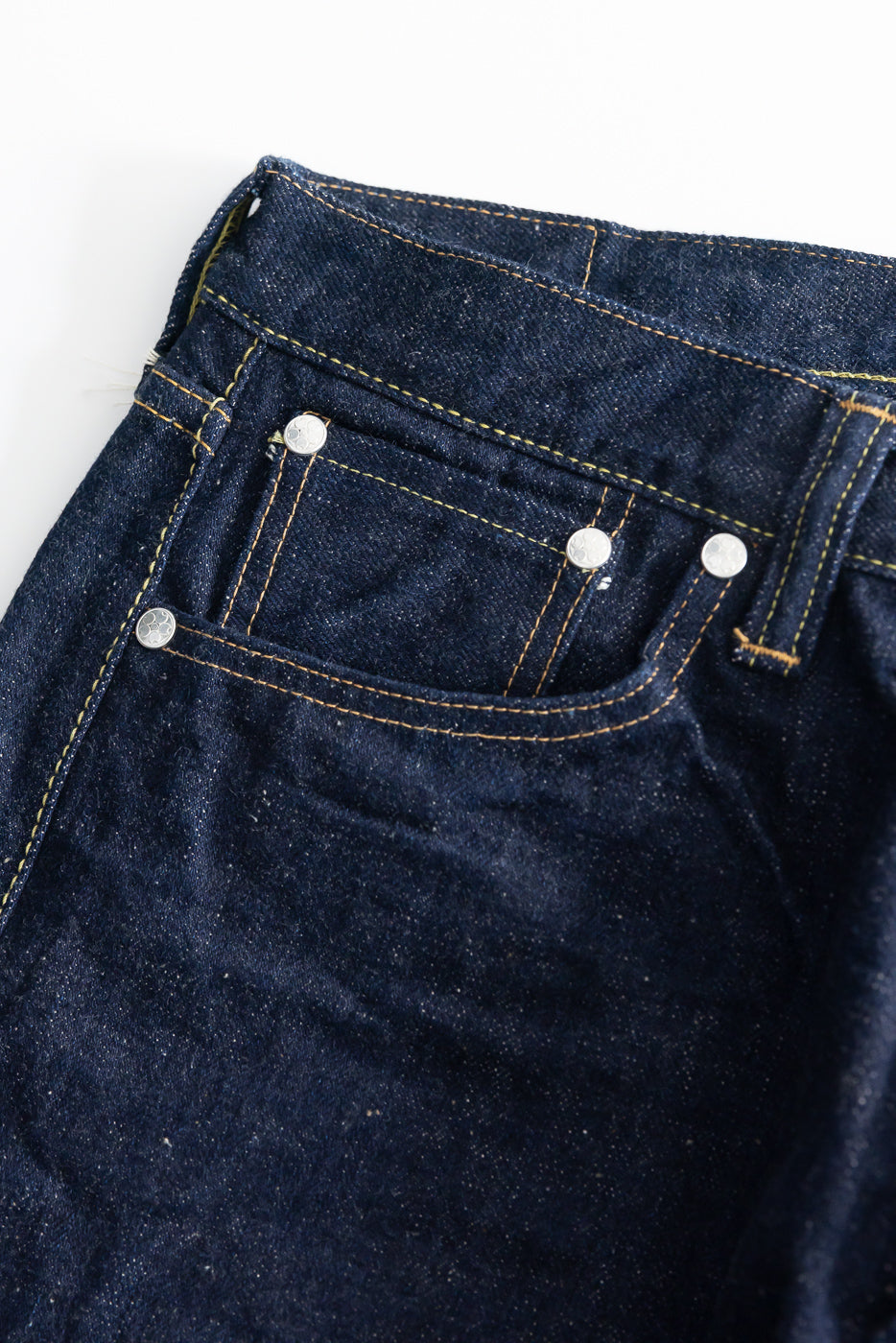 Close-up of front left pocket and coin pocket on dark indigo selvedge jeans, visible rivets, yellow contrast stitching.