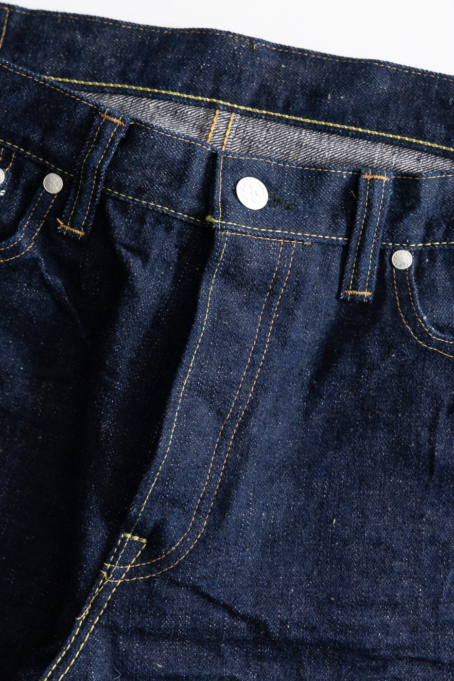 Close-up of front waist and fly area of selvedge jeans, showing denim texture, stitching, metal rivets, and button details.