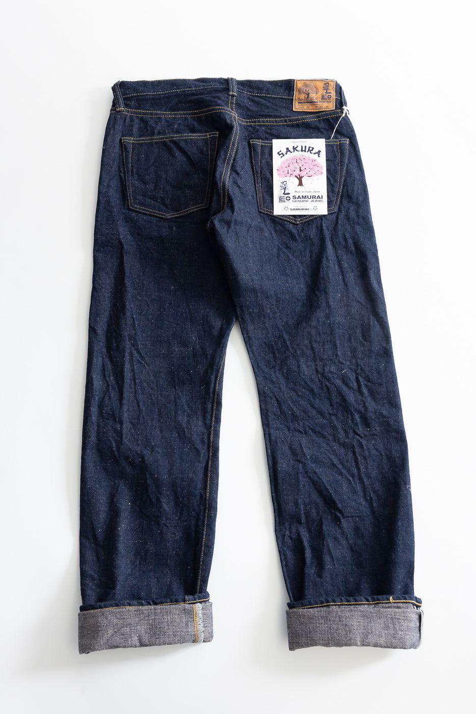 Flat lay of selvedge jeans back side, showing rear pockets, leather patch, and sakura tag detail.