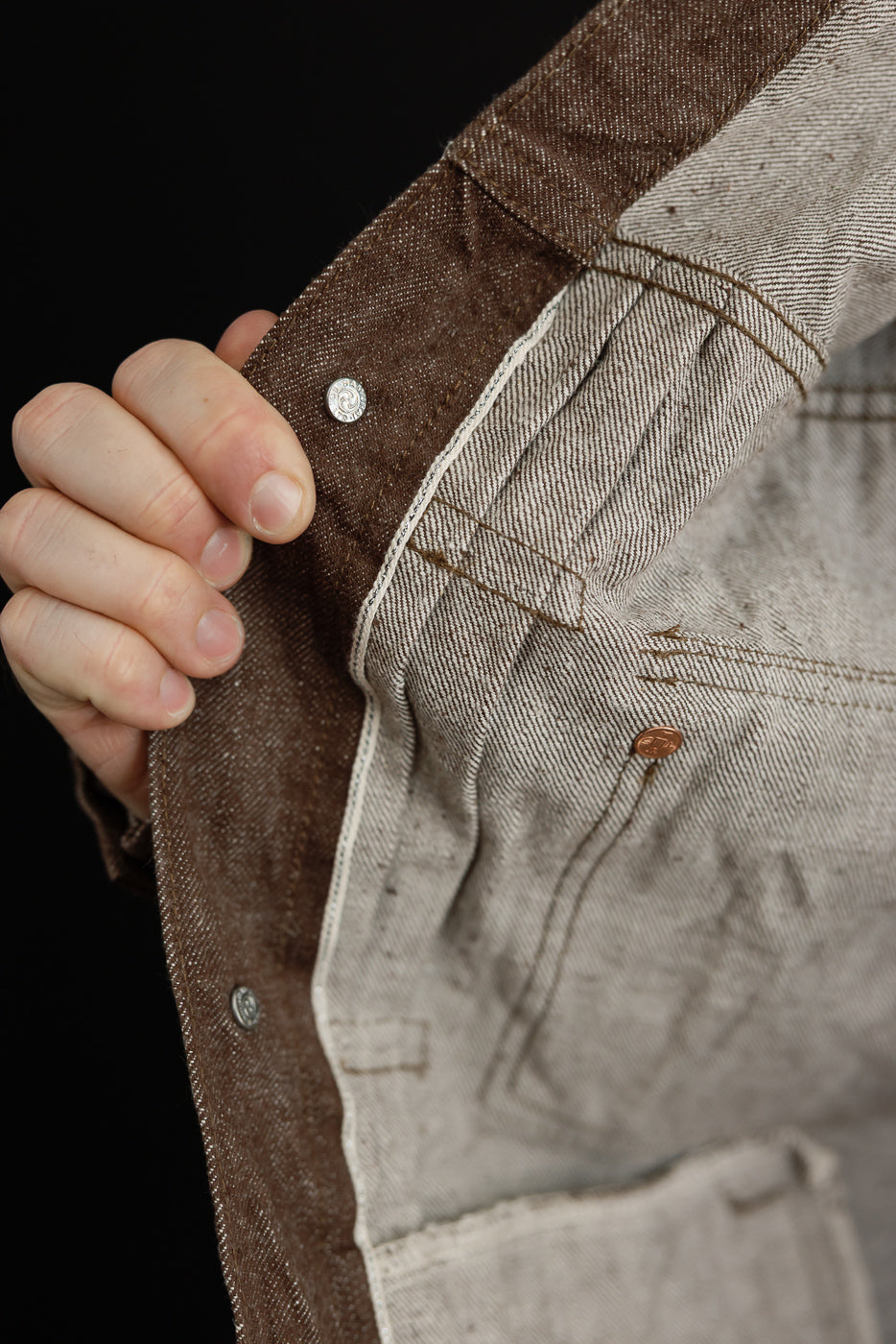 Close-up of men's selvedge denim jacket front, showing brown fabric, silver buttons, and stitch details