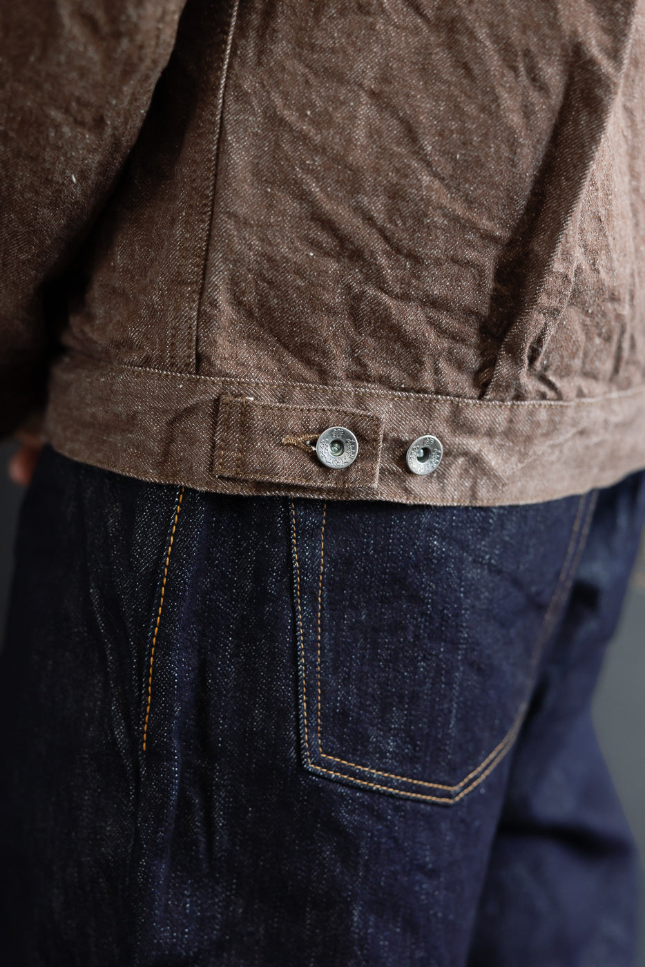 Close-up side view of brown denim jacket hem with silver buttons, worn by male model, paired with dark selvedge jeans
