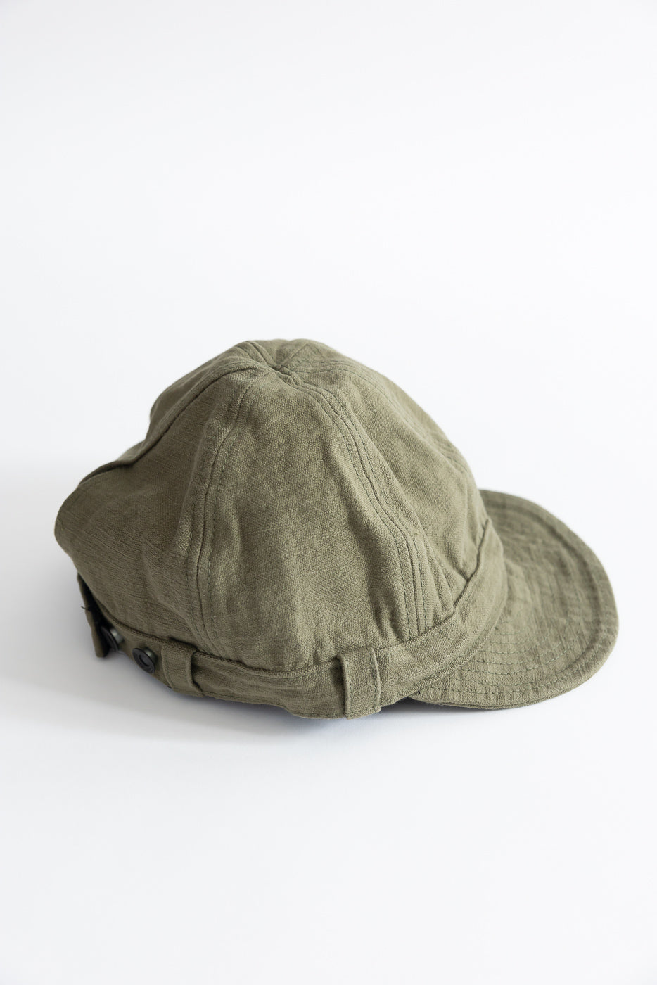 Close-up flat lay of olive green workman’s cap, showing top and side details, textured fabric, and button accents.