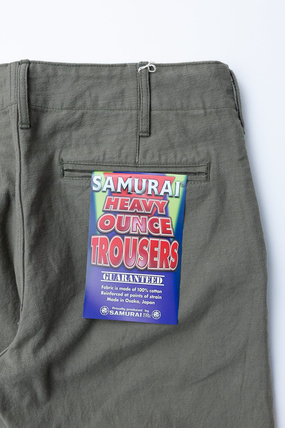 Front close-up of olive Samurai Heavy Ounce Trousers, showcasing tag details and 100% cotton fabric texture.