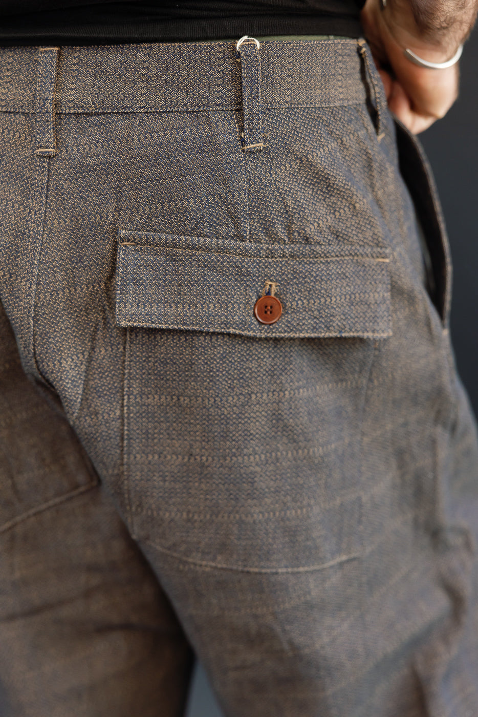 Close-up back view of men's textured selvedge trousers, showing buttoned pocket, waistband, and fabric detail on athletic build model