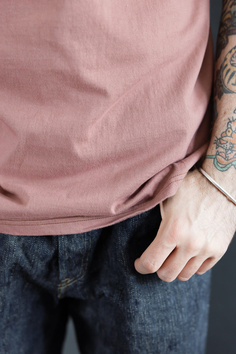 Close-up of male model's torso in dusty rose tee, showcasing hem and fabric texture, paired with selvedge denim
