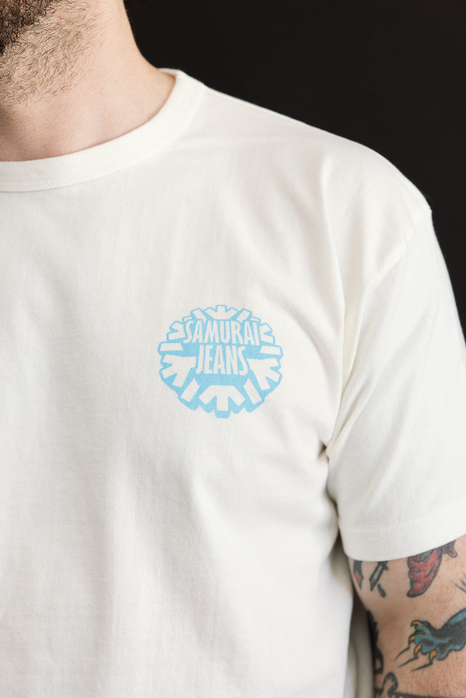 Close-up front view of male model wearing off-white tee with light blue Samurai Jeans logo, crew neck, short sleeves