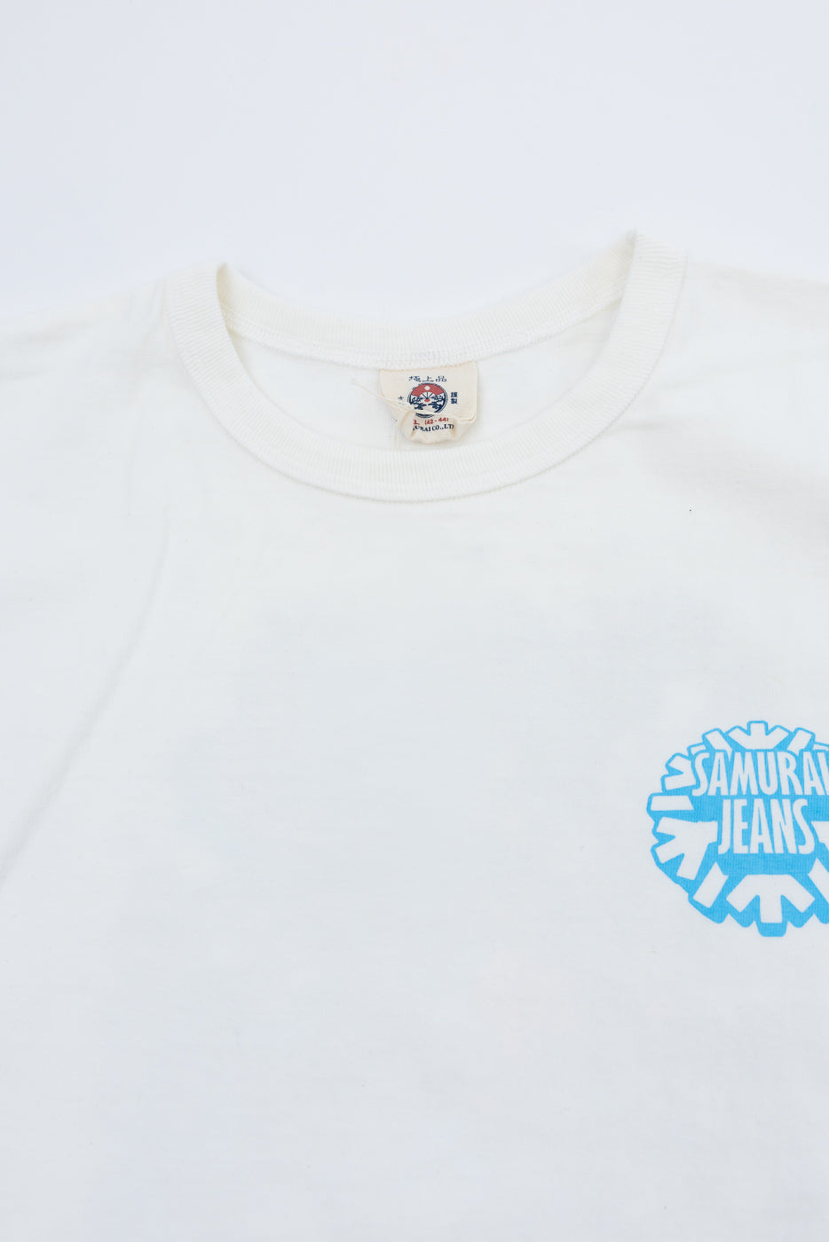 Close-up front view of white t-shirt with blue Samurai Jeans logo, ribbed collar, and brand tag detail