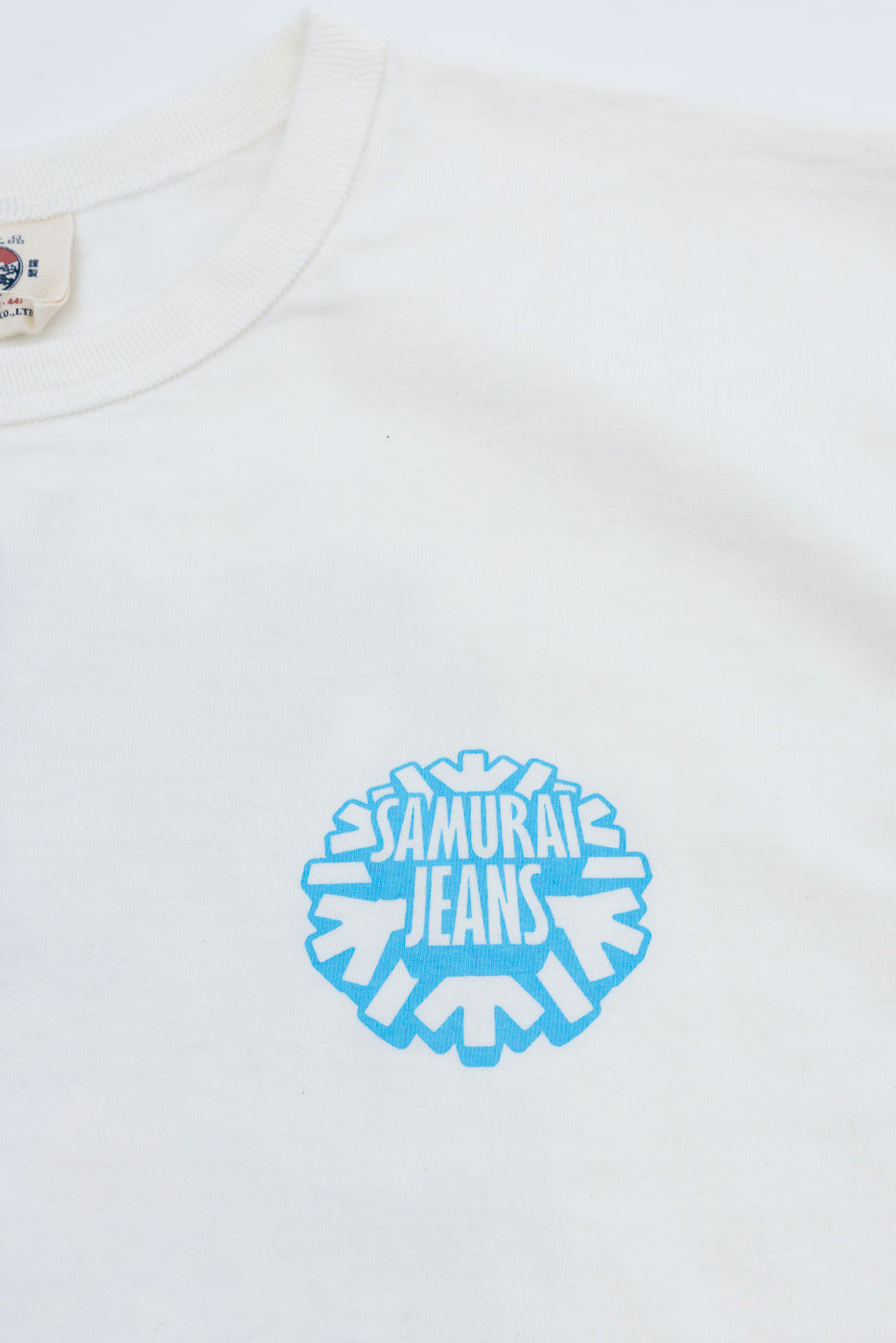 Close-up front of white tee with blue Samurai Jeans logo, tag visible on collar