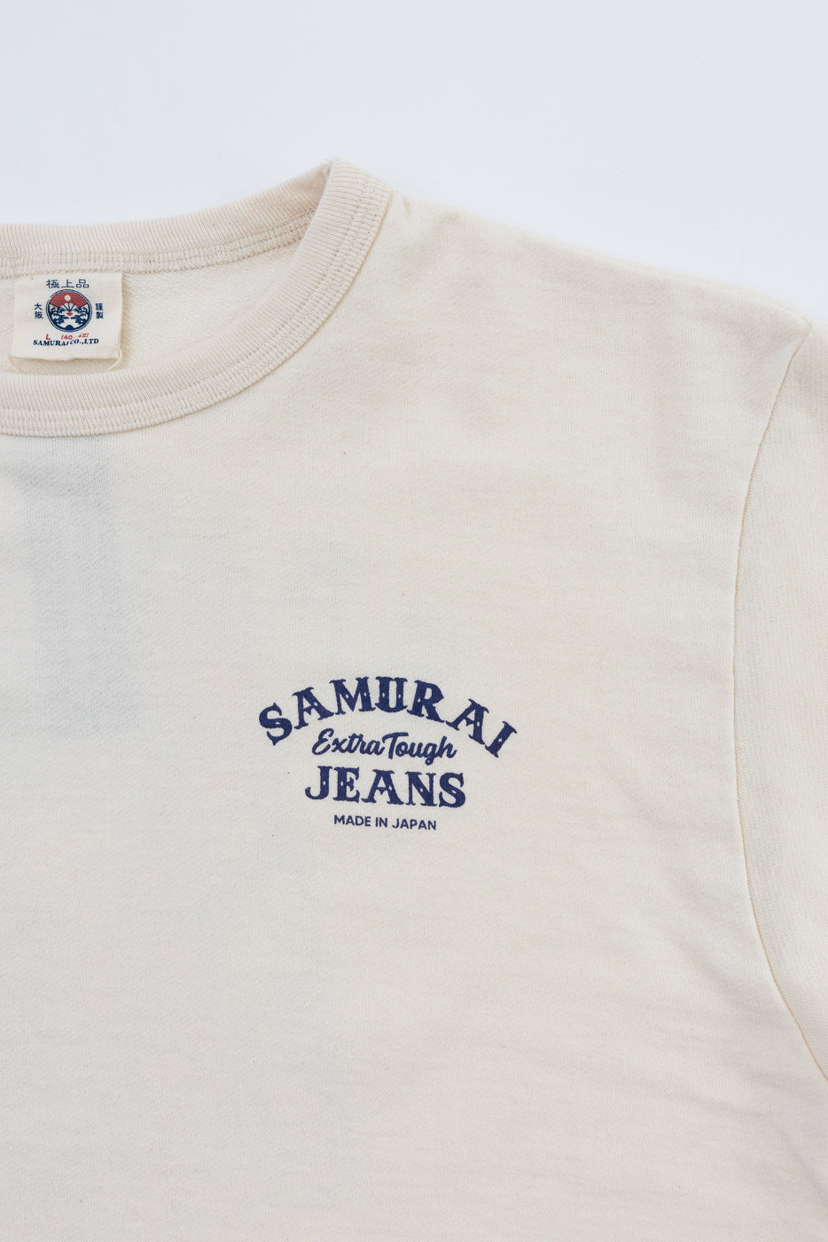 Close-up front of cream Samurai Jeans tee, showing chest logo, tag, and ribbed crew neck