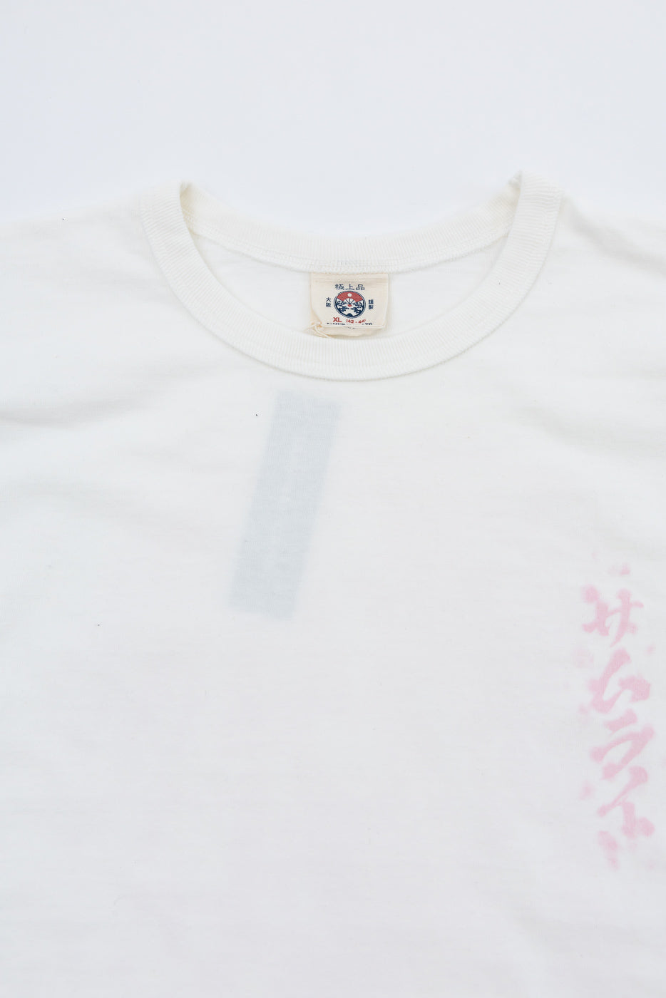 Close-up front view of white tee showing collar, tag with Japanese text, and faint pink graphic detail.