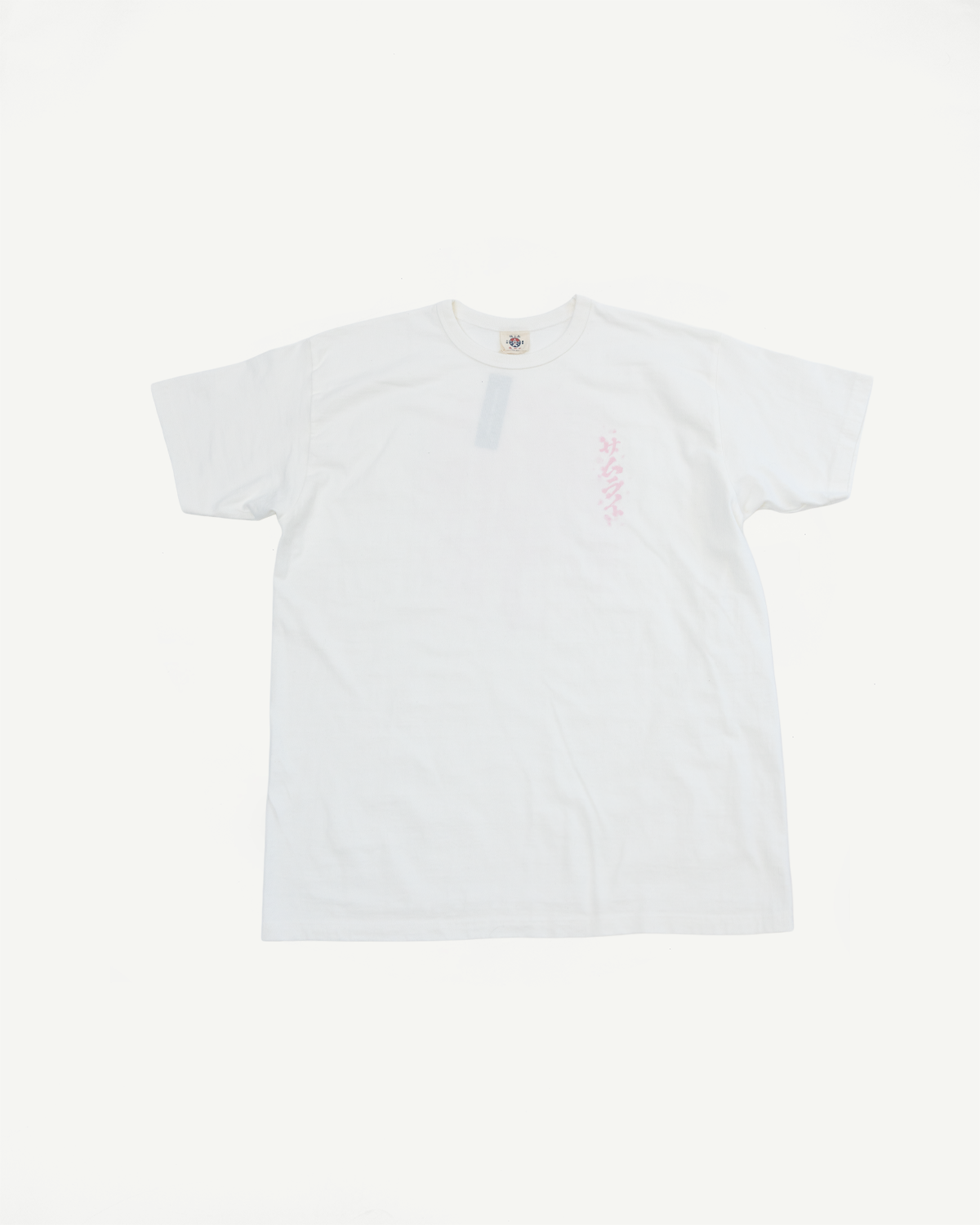 White crewneck t-shirt flat lay, front view, pink graphic on left chest, tag visible at collar
