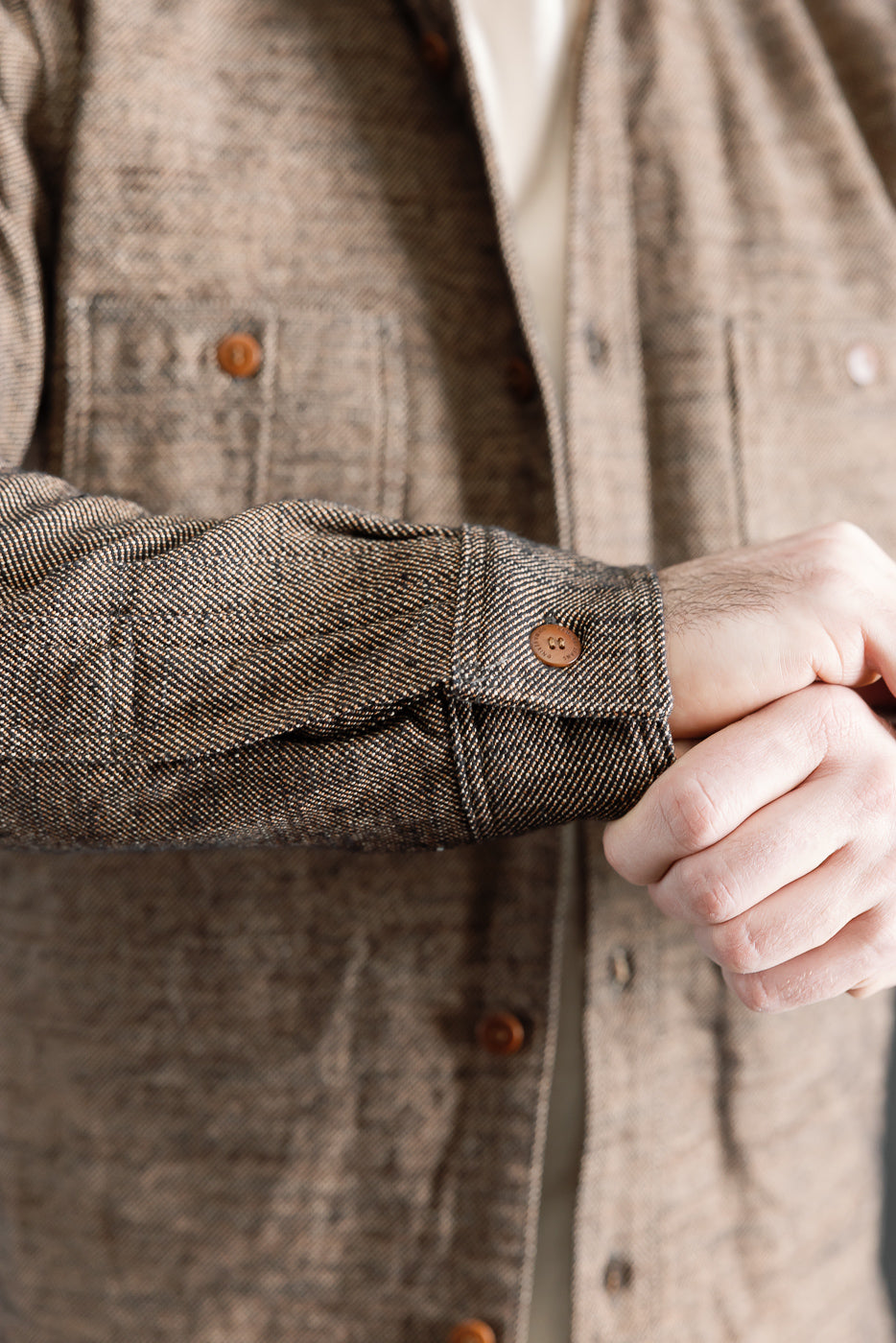 Close-up of male model's right sleeve cuff and button on brown flannel shirt, showing front detail and fabric texture, Dant contemporary casual apparel.