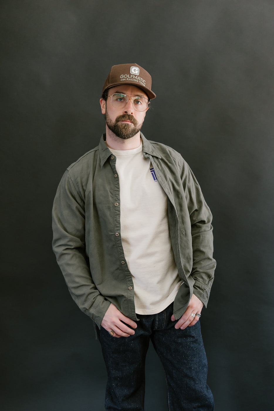 Man in olive green Tropical Weather Dobby shirt with clear-lens glasses and baseball cap, showcasing the shirt's military-inspired, textured design and casual style.