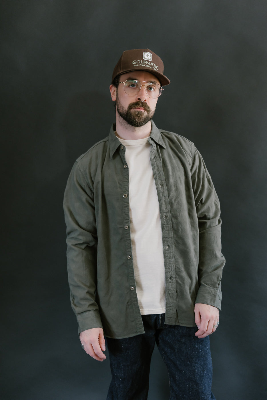 Man wearing the SSS25-ATW Tropical Weather Dobby Shirt in olive green, styled with a brown cap and jeans, showcasing its relaxed fit and textured design.