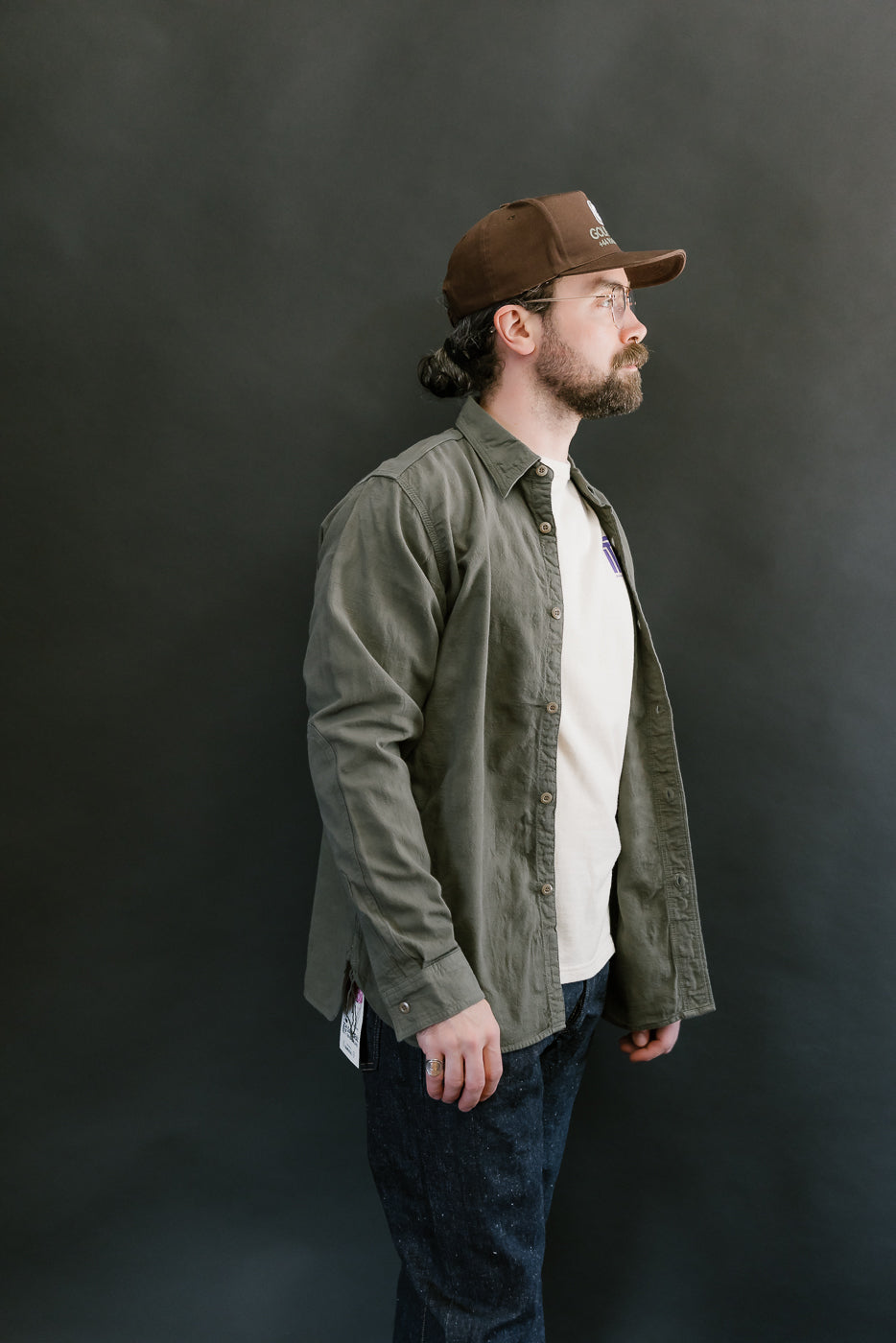 Man models SSS25-ATW Tropical Weather Dobby Shirt in olive green, showcasing its open style over a T-shirt, paired with dark denim jeans.