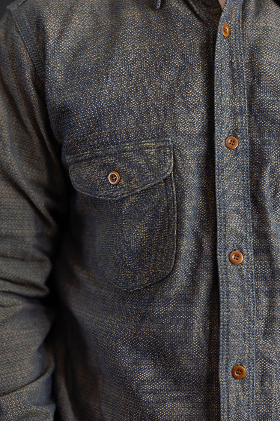 Close-up front view of men's denim shirt with chest pocket, brown buttons, and textured fabric, worn by male model