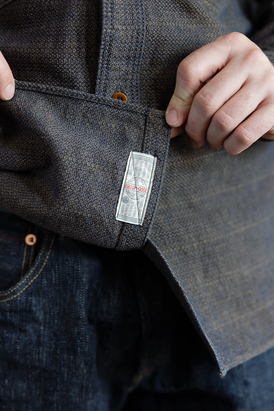 Close-up of male model's left side showing denim shirt pocket with tag, button, and selvedge fabric texture