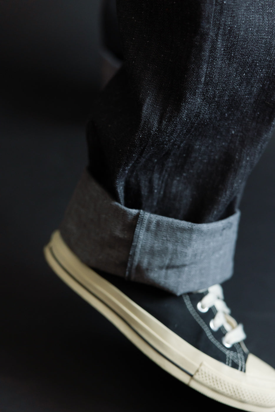 Close-up of male model's cuffed selvedge denim hem, showing raw edge texture and black sneaker pairing